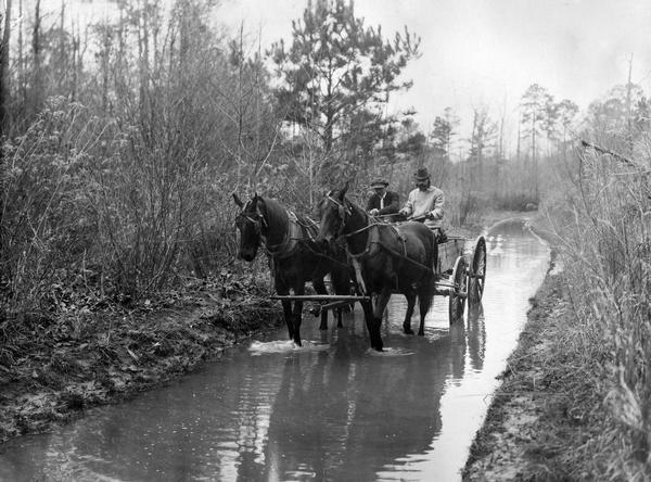 Two men traveling in a horse-drawn wagon on a flooded rural road. Original caption reads: "Example of road building in Lamar County on the Vernon-Columbus road. For more than one-quarter of a mile the main road is made a drainage ditch for the surrounding territory. As the land on either side is not very valuable, being grown up to brush and weeds, there is no very good reason why the road should be at the bottom of the lake, although it seems to have been constructed that way. C.C. Hudson, in wagon, the trip from W.O. Pennington's to Mt. Pleasant School, February 13, 1915."