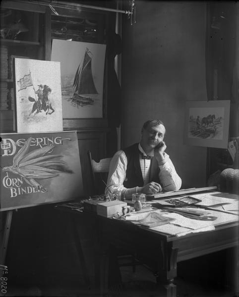 International Harvester advertising artist sitting at his desk in a studio office with a wistful look on his face. Graphics from Deering brand advertisements are propped up behind him. The photograph was likely taken at the company's McCormick Works.