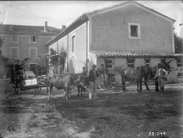 Farmers standing in a farm yard with a McCormick binder pulled by oxen and a horse-drawn mower. The farm appears to be in France.
