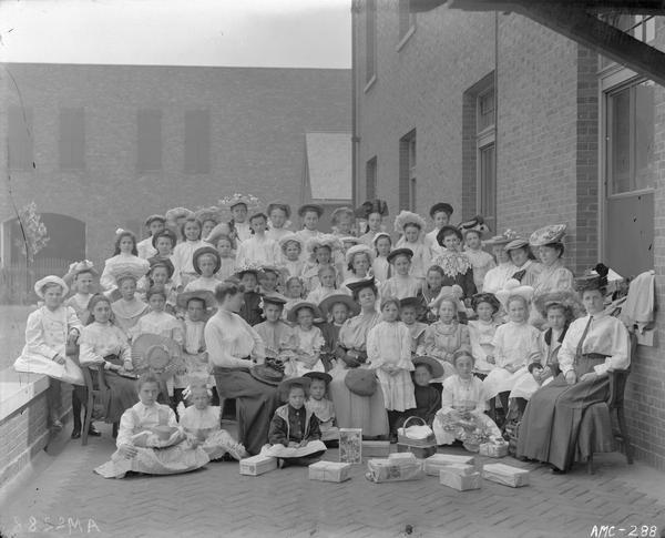 Outdoor group portrait of women and young girls posing in dresses and flowered hats in an urban area, perhaps near the McCormick Works Club House. There appear to be gifts, some wrapped in newspaper, laid out on the ground in front of the group.