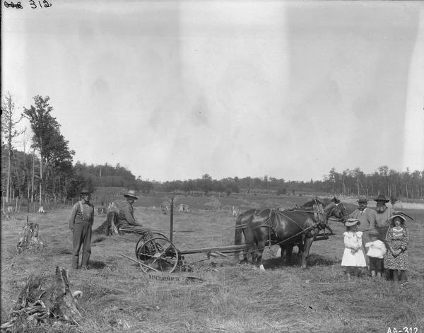 Four men and three children standing in a stump-strewn field with a horse-drawn McCormick mower.