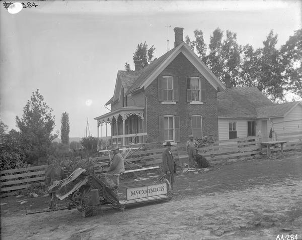 Men, young boys, and a dog standing in front of a brick farmhouse with a horse-drawn McCormick binder.