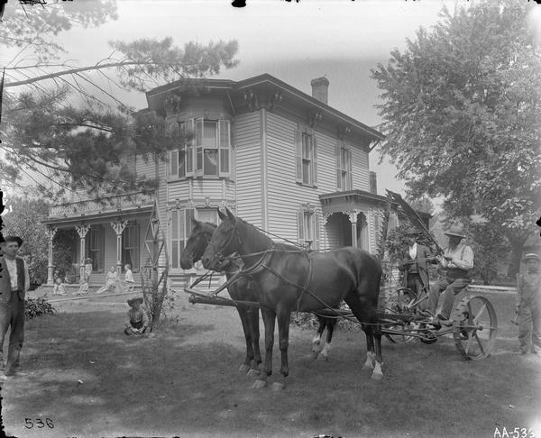 Farm family posing around a house and yard, with the patriarch sitting in the seat of a mower behind two horses.