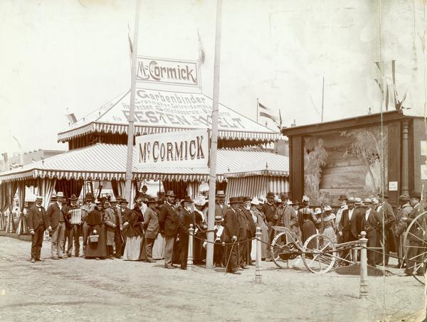 A crowd of people gathered at the entrance to a German exhibition of McCormick agricultural equipment. To the right is a small theatrical stage depicting a farmer harvesting grain and a McCormick mower.