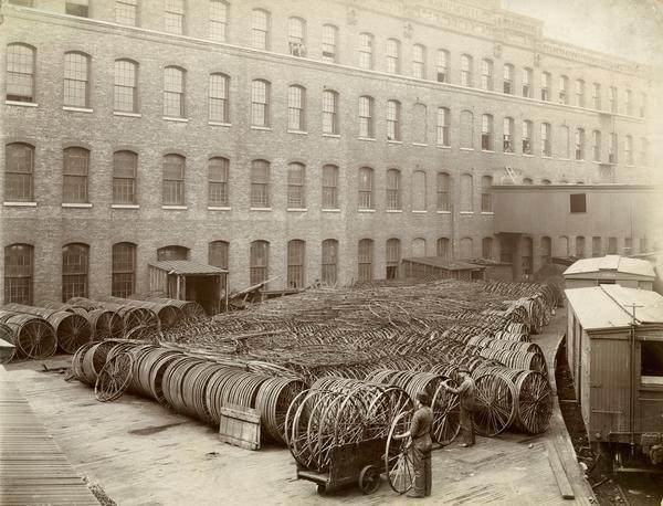 Two workers among hundreds of hay rake wheels stacked on a shipping platform next to rail cars at the McCormick Reaper Works. The factory was owned by the McCormick Harvesting Machine Company until 1902 when it became the International Harvester Company's "McCormick Works". The factory was located at Blue Island and Western Avenues in the Chicago subdivision called "Canalport". It was closed in 1961.