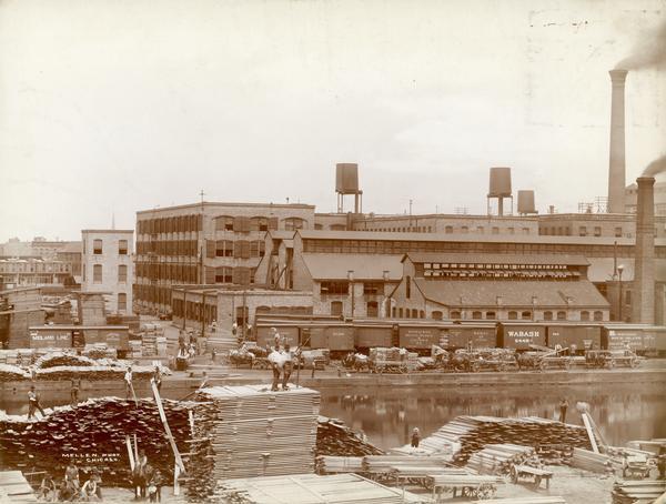 Workers loading and unloading wood at the wood stock yard of the McCormick Reaper Works. Men are working on both sides of the Chicago River. Rail cars and wagons piled with lumber are parked on the far side of the river. The factory was owned by the McCormick Harvesting Machine company before 1902. In 1902 it became the McCormick Works of the International Harvester Company. The factory was located at Blue Island and Western Avenues in the Chicago subdivision called "Canalport". It was closed in 1961.