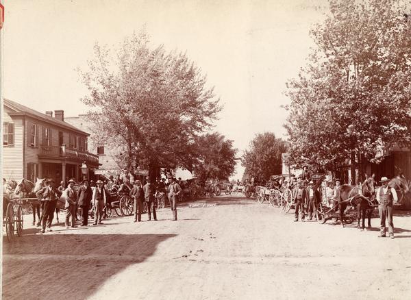 Farmers lined up with horse-drawn wagons loaded with newly delivered McCormick farm machines on the main street of a rural town. The wagons are adorned with "McCormick" signs. "McCormick Days" were events organized by local dealers or agents to publicize the delivery of machines to customers.