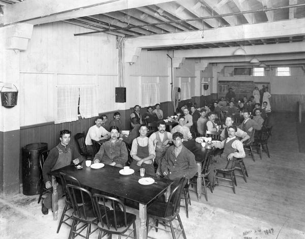 Factory workers sitting at tables in a cafeteria at International Harvester's Osborne Works. The factory was owned by the D.M. Osborne Company until 1903, when it was purchased by International Harvester. The factory was later known within the company as the "Auburn Works." It was located at 5 Pulaski Street.