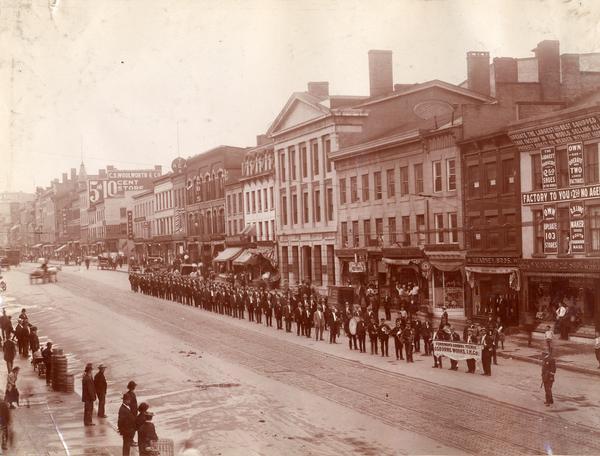 Elevated view of men following a band in a parade through downtown Auburn for the Osborne Works foremen's annual picnic. Spectators are standing on the sidewalks of the city street, and storefronts are in the background. The Osborne Works was owned by the D.M. Osborne Company until 1903, when it was purchased by International Harvester. The factory was later known within the company as the "Auburn Works". It was located at 5 Pulaski Street.