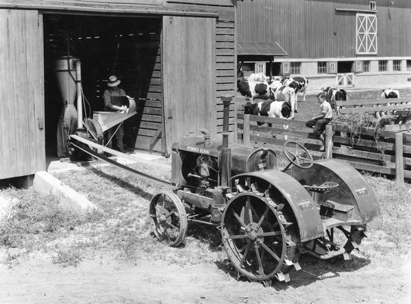 Man using a McCormick-Deering W-30 tractor to run a belt-driven hammer mill as a boy is watching from a fence. The hammer mill is likely an International No. 1B (continued by the No. 10). A barn and dairy cows are in the background.