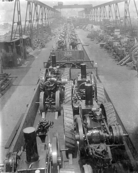 Elevated view of Mogul tractors loaded onto railroad cars outside International Harvester's Tractor Works. A variety of parts, engines, implements, and tractors are parked or stacked along both sides of the rail line.