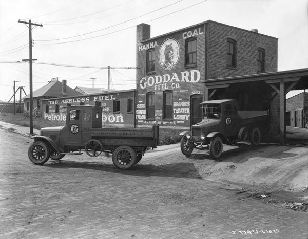 Two International delivery trucks leaving premises of the Goddard Fuel Company. The company's building and trucks carry advertising  for "Hanna Coal" which features an illustration of an African American woman.