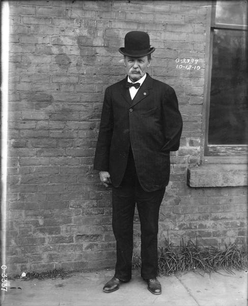 One-armed factory worker in a suit and bowler hat posing against a brick wall. The man likely worked at International Harvester's Osborne Works.