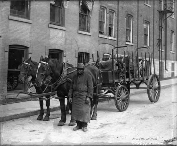 African American wagon driver standing with a horse-drawn wagon parked on a city street. The man is likely an employee of International Harvester, possibly at the company's Osborne Works. The wagon bears the company's name along the bed.
