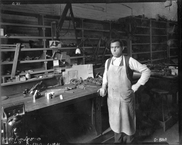 Worker standing in front of a work bench covered with tools, parts and parts drawings at International Harvester's Osborne Works (later known as "Auburn Works"). Notices promoting Charles F. Hawelka and Clarence Heieck for "trustee" are pinned above the desk. The Osborne factory was originally owned and operated by the D.M. Osborne Company.