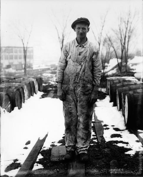 Factory worker in soiled clothing standing among wooden barrels in a snow-covered factory yard. The man most likely worked at International Harvester's Osborne Works.
