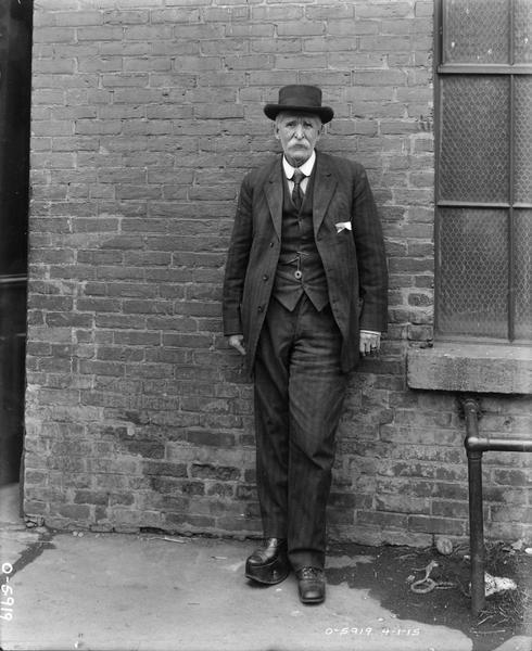 Old man wearing a suit and hat with his back against a brick wall. The man is likely an employee at International Harvester's Osborne Works. One of his legs is shorter than the other and he is wearing a corrective shoe.