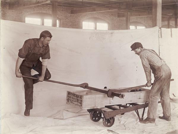 Two factory workers attempting to twist an implement part with large wrenches at International Harvester's McCormick Works. The part is resting on a crate for the McCormick no. 6 binder. The men are working in front of a white sheet that is serving as a backdrop for the photographer.