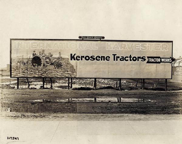 Billboard advertising International Harvester kerosene tractors along muddy dirt road outside International Harvester's Tractor Works. The billboard was created by Thos. Cusack Co. and shows a Mogul 10-20(?) HP tractor with plow. The factory was located at 2600 West 31st Boulevard.