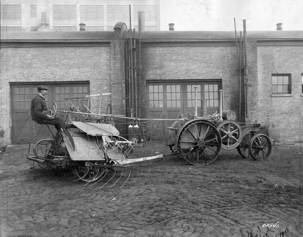 Man operating a Deering New Ideal binder pulled by a Mogul 8-16 HP tractor outside an International Harvester factory. The man is sitting in the seat of the binder and is holding an extended steering wheel attached to the tractor.