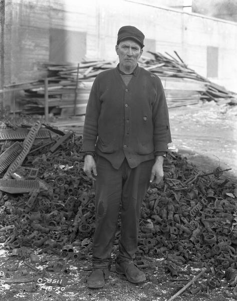 Older factory worker standing in front of a pile of scrap parts in a factory yard, most likely at International Harvester's Osborne Works.