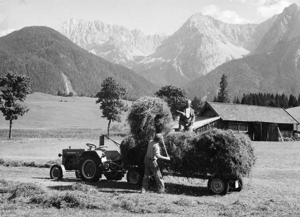 German farmers baling hay in southern Germany's Alpine country. The men are using an International D-214 tractor with mower and wagon. Original caption reads: "High in southern Germany's Alpine country, mechanized farm equipment is rare but modern engineering produces machinery like this McCormick International tractor and mower that operate efficiently even at high altitudes. The expanding economy of the big city is spreading to remote areas like this and industrious Germans are quick to adapt the latest technology to their specific interest."