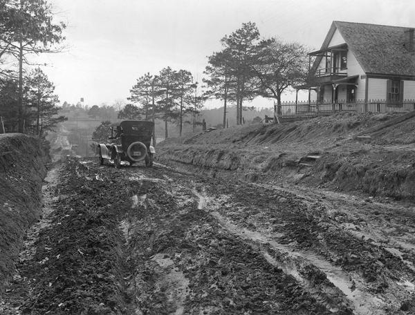 Man driving an automobile down a muddy dirt road. Caption on back reads: "Grading road with surfacing." The photograph was taken for International Harvester's Agricultural Extension Department to document the condition of rural roads in the South.