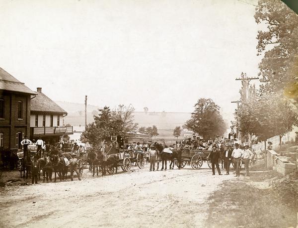 Families and citizens with horse-drawn wagons gathered in a small town for delivery of McCormick farm machines from General Agent Daniel Klinedinst. Horses and wagons are adorned with "McCormick" signs and the advertising poster "Back from the War". The town was in York County.