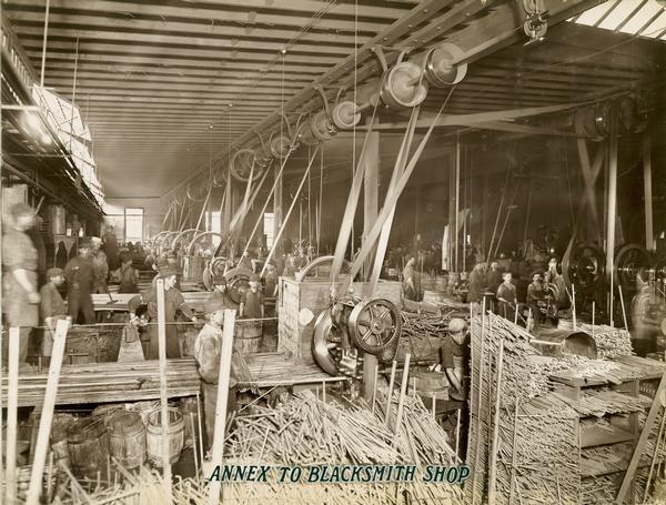 Men and boys working with steel rods and belt-driven machinery to make bolts inside an annex to the blacksmith shop at the McCormick Reaper Works. The factory was owned by the McCormick Harvesting Machine company before 1902. In 1902 it became the McCormick Works of the International Harvester Company. The factory was located at Blue Island and Western Avenues in the Chicago subdivision called "Canalport." It was closed in 1961.