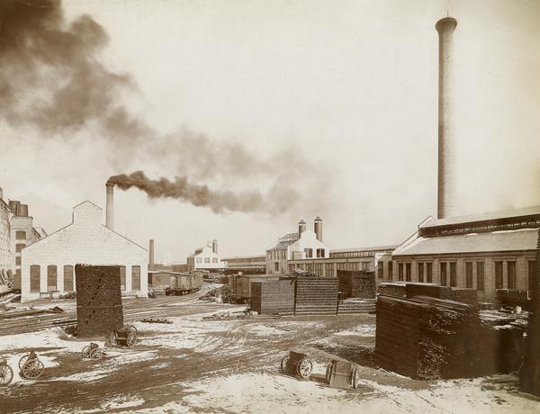 Small carts, stacks of lumber, and rail cars in a snow-covered yard at the McCormick Reaper Works. The original caption reads: "Foundry #28, Wheel Room, Blacksmith Shop #2". The factory was owned by the McCormick Harvesting Machine company before 1902. In 1902 it became the McCormick Works of the International Harvester Company. The factory was located at Blue Island and Western Avenues in the Chicago subdivision called "Canalport." It was closed in 1961.