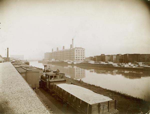 McCormick Reaper Works and wood shop stock yard as seen from across a canal. In the foreground is a rail yard with a small engine and rail cars. The factory was owned by the McCormick Harvesting Machine company before 1902. In 1902 it became the McCormick Works of the International Harvester Company. The factory was located at Blue Island and Western Avenues in the Chicago subdivision called "Canalport". It was closed in 1961.
