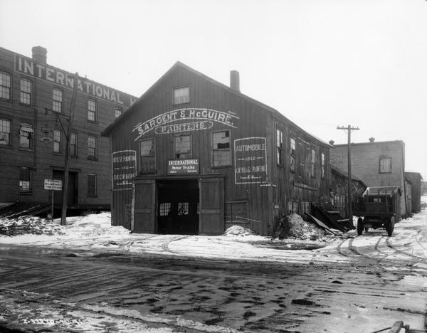 International motor truck dealership and branch house(?) buildings. A wooden building has a small "International Motor Trucks" sign over the door. However, the name "Sargent & McGuire, Painters," is on the building itself. A large brick building in the background appears to be an International Harvester branch house.