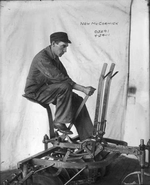 Man sitting on a "new" McCormick riding disc harrow, probably in an International Harvester factory. He is sitting in front of a  makeshift white backdrop.