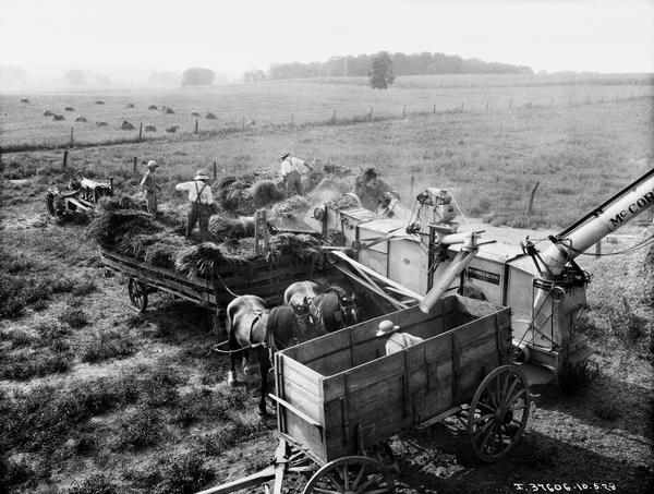 Elevated view of farmers threshing grain in a field with a belt-driven McCormick all-steel thresher run by a Farmall tractor. One person is working inside of a high-walled wooden wagon, and three men are standing on top of horse-drawn hay carts.