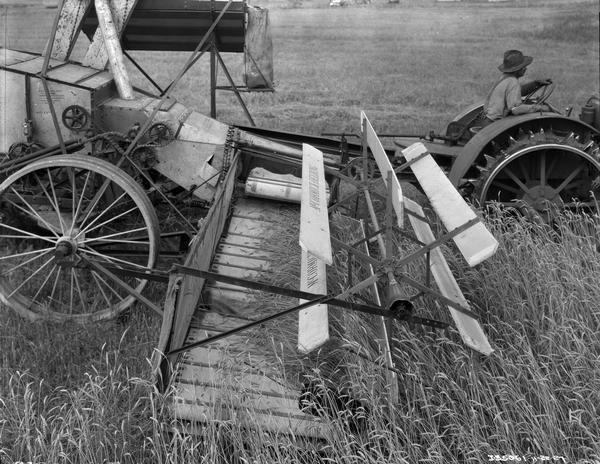 View of right side of a McCormick-Deering combine (harvester-thresher) pulled by a Farmall tractor in a field. A man wearing a hat is driving the tractor.