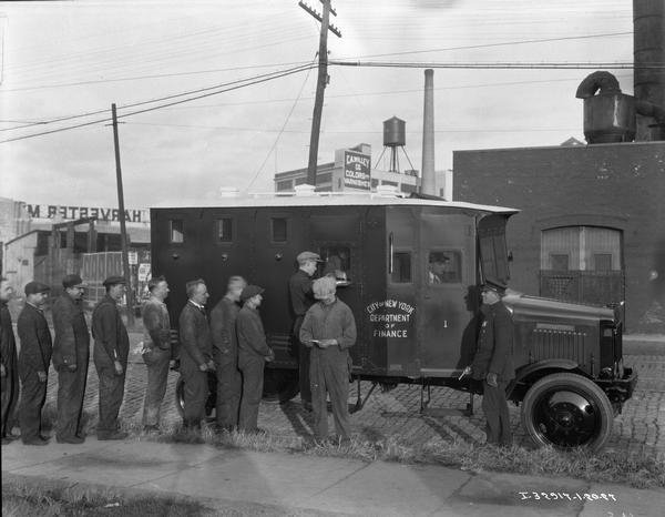 Men receiving paychecks from armored trucks parked outdoors at an IH plant. Signs on the trucks read: "Department of Finance, City of New York." A police officer standing near the truck on the right is holding a handgun.