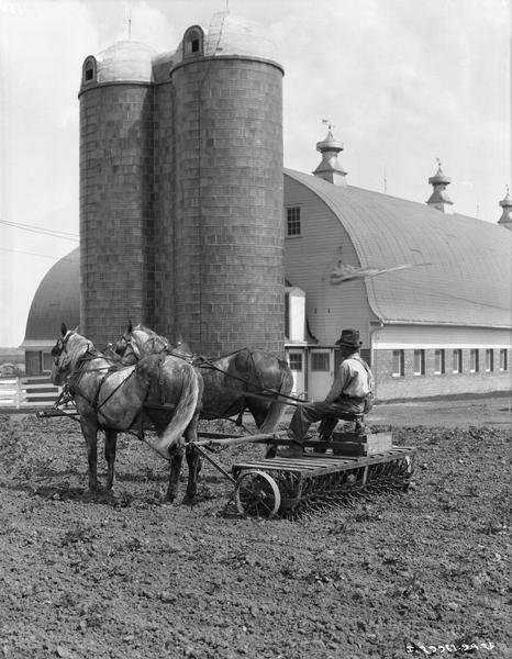 Farmer cultivating a field with a horse-drawn McCormick rotary hoe in front of silos and farm buildings.