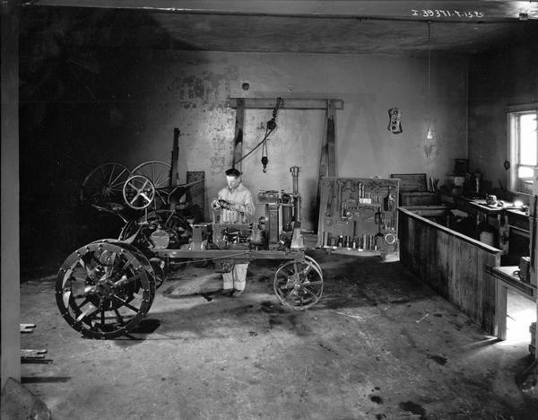 Mechanic inspecting a part from a Farmall tractor in a repair shop. A tool rack is in the background. The shop is likely part of an International Harvester dealership.