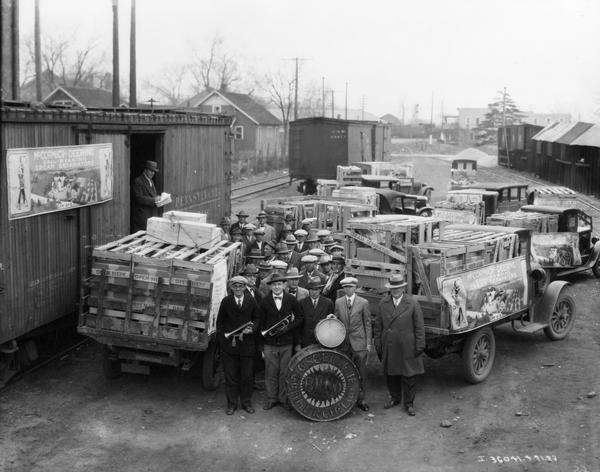 Elevated view of the Commercial Club Band posing at a rail yard with a newly arrived shipment of McCormick-Deering ball-bearing cream separators. The cream separators have been unloaded in crates onto International trucks. Large advertising posters for McCormick-Deering cream separators are mounted on the side of the rail car and on one of the trucks. The event likely was arranged by an International Harvester dealer as a public relations event.