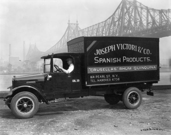 Driver in an International delivery truck parked along the waterfront with the Queensboro Bridge in the background. The truck was owned by Joseph Victori & Co., purveyors of "Spanish Products." The rest of the sign painted on the side of the truck bed reads: "'Crusellas' Rhum Quinquina."