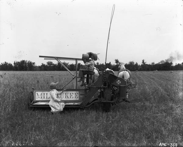 Young girl and boy posing with a horse-drawn Milwaukee grain binder in a field. The boy is drinking from a jug.