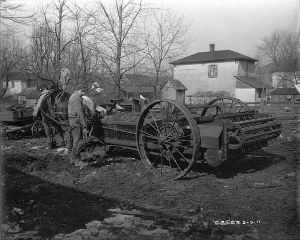 Farmer shoveling manure into the box of a horse-drawn International New Low manure spreader in a farm yard.