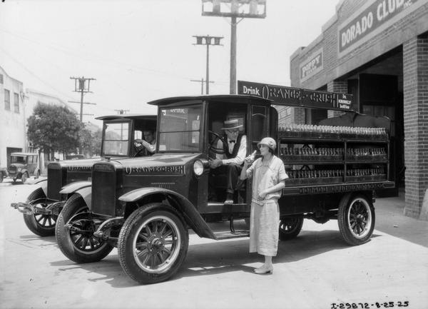 Young woman drinking a bottle of Orange Crush soda in front of two International Model S Orange Crush delivery trucks parked along a storefront. A driver looks on from the seat of his delivery truck.