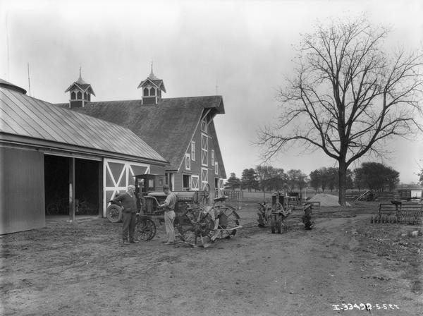 Two men standing next to a Farmall Regular tractor in a farm yard outside a barn. One man may be an International Harvester dealer providing service. An International truck is parked in the background.