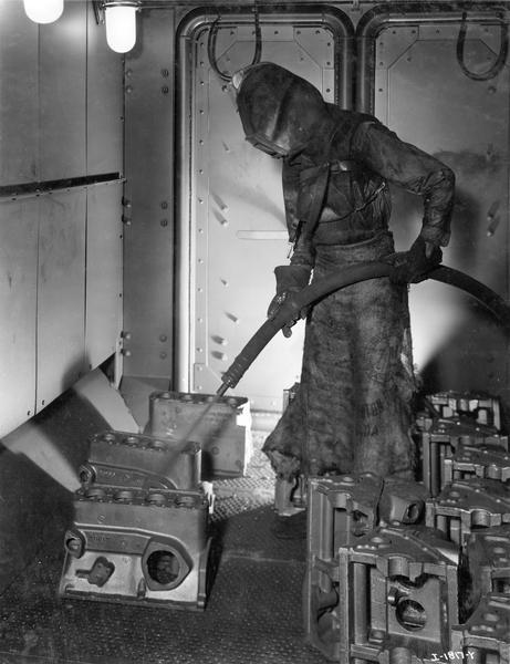 Factory worker in a protective suit, mask, and gloves cleaning engine blocks in the foundry sand blast booth at International Harvester's Tractor Works.