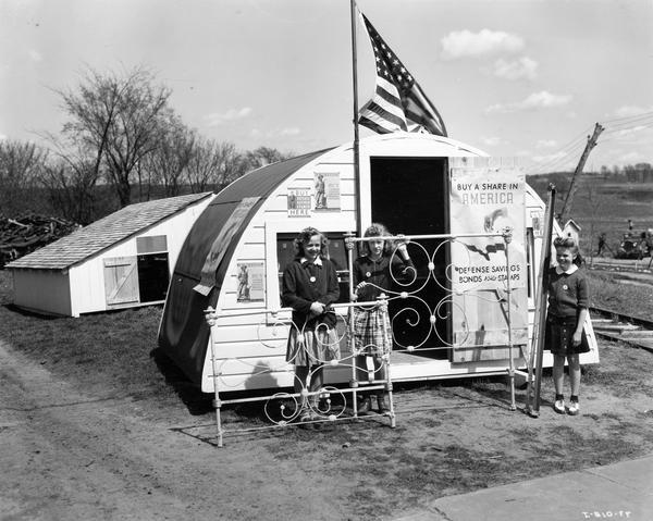 Three young girls, (L to R): Beverly Peterson, Virginia Gullickson, and Joane Aaby, with an old iron bed donated for a WW II scrap drive during MacArthur Week activities. The girls are standing at the entrance to the town scrap pile in front of a booth selling war bonds and stamps. 182 farmers participated in the drive that brought in over 105 tons of scrap metal to this town of less than 500.