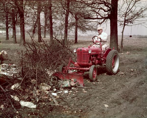 Color photograph of a worker clearing trash and other debris using an International Cub Lo-Boy tractor outfitted with a front-mounted IH hydraulic grading blade.