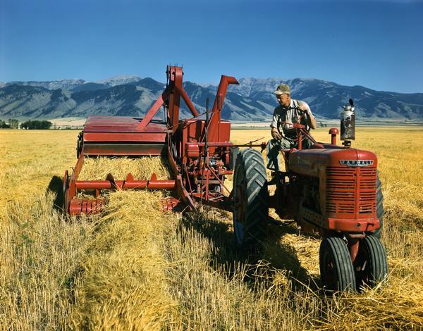 Farmer on a McCormick-Deering Farmall H tractor with no. 64 combine (harvester-thresher) set against a mountainous backdrop.
