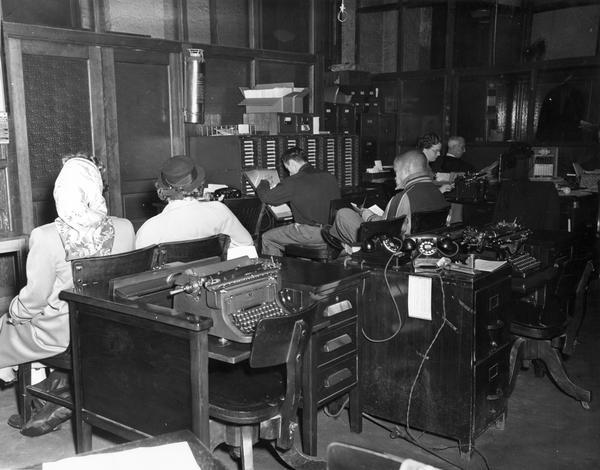 Men and women wait in an International Harvester company office among desks, typewriters, and other office equipment.
