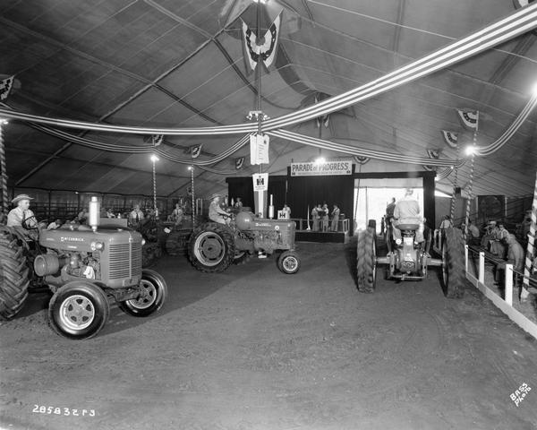 Men in cowboy hats driving International Harvester tractors through an exhibition tent as part of the "Parade of Progress" at the Indiana State Fair as part of the Fast Hitch Square Dance. In the background a four-piece band plays while the announcer calls the square dance. Men and women view event from side bleachers. Tractors include the McCormick Super WD-6, the Farmall M, and a few crawler tractors.